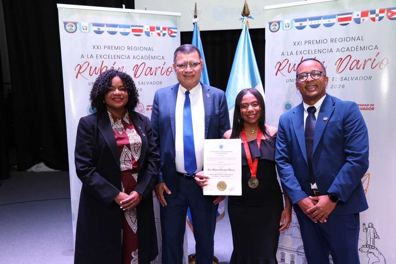 Enny Virginia Lantigua, con su medalla y su certificado, acompañada de tres autoridades universitarias frente a los telones del premio en San Salvador